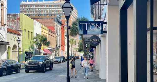 A bustling street scene with shops, pedestrians, and parked cars under a clear blue sky. Palm trees line the sidewalk.