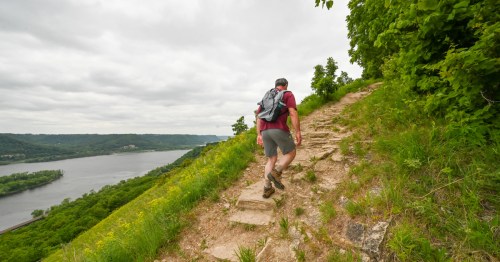 A hiker ascends a rocky trail beside a river, surrounded by lush greenery and cloudy skies.