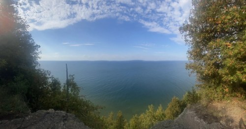 Panoramic view of a calm lake surrounded by trees under a partly cloudy sky.