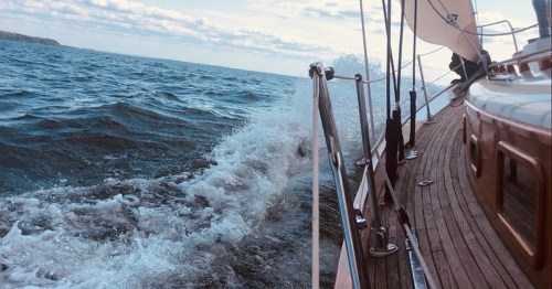 A sailboat glides through choppy waters, with waves splashing against the side and a cloudy sky above.