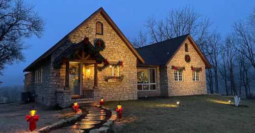 A cozy stone house decorated with wreaths and lights, surrounded by trees, illuminated at dusk.