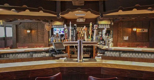 A wooden bar with a polished countertop, shelves of liquor, and a beer tap, set in a cozy, rustic pub interior.