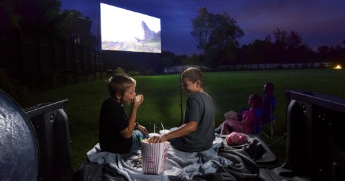 Two boys sit on a blanket in a truck bed, enjoying popcorn while watching a movie on an outdoor screen at dusk.