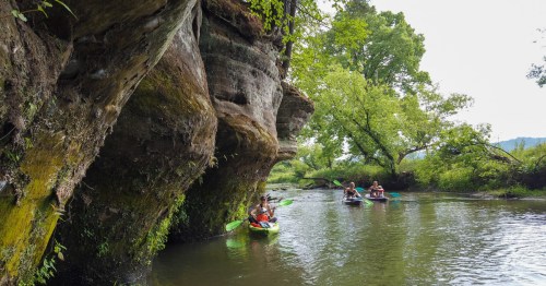 Three kayakers paddle along a serene river, flanked by lush greenery and towering rock formations.