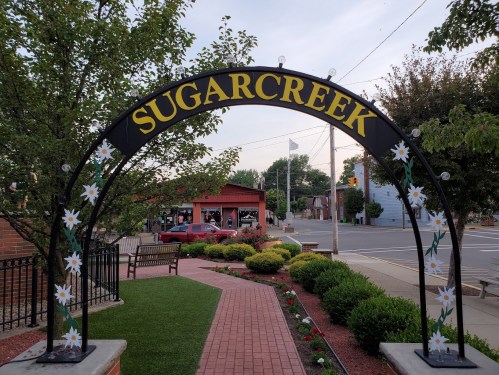 Archway sign reading "SUGAR CREEK" adorned with flowers, leading into a landscaped area with benches and greenery.