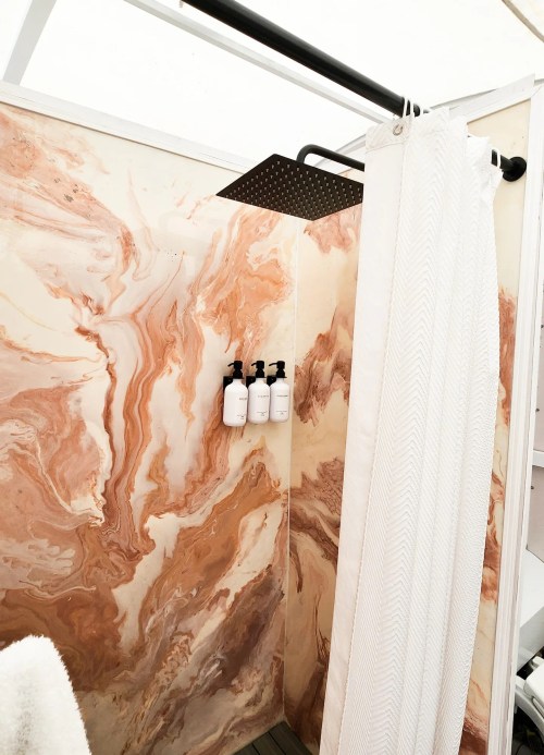 A modern shower with a marble-patterned wall, a rainfall showerhead, and three white bottles on a shelf.