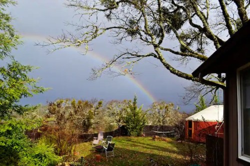 A rainbow arcs over a garden with trees, a wooden fence, and a yurt, under a cloudy sky.