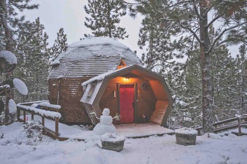 A unique, snow-covered dome-shaped cabin with a red door, surrounded by pine trees and a snowman in the foreground.