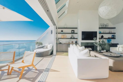 Bright, modern living room with ocean views, featuring a white sofa, wooden chairs, and large glass doors.