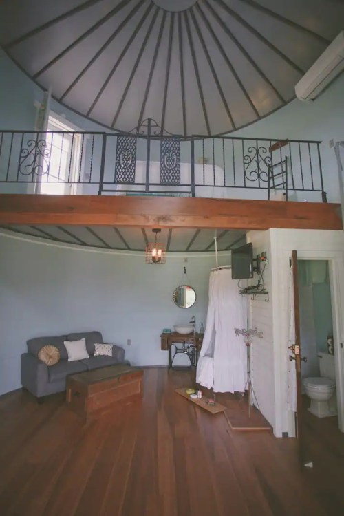Cozy room with a circular ceiling, wooden floors, a lofted bed, and a sitting area featuring a sofa and decorative elements.