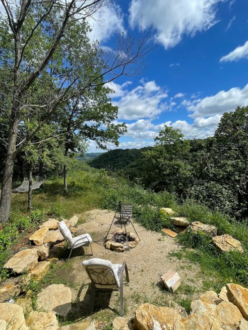 A serene outdoor scene featuring a fire pit surrounded by chairs, with lush greenery and a blue sky in the background.
