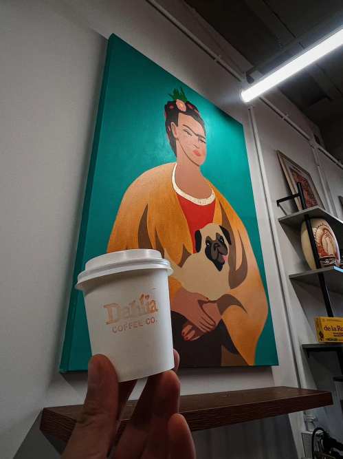 A hand holds a coffee cup in front of a colorful painting of a woman with a dog.