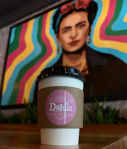 A coffee cup with a pink label in front of a colorful mural featuring a woman's portrait.