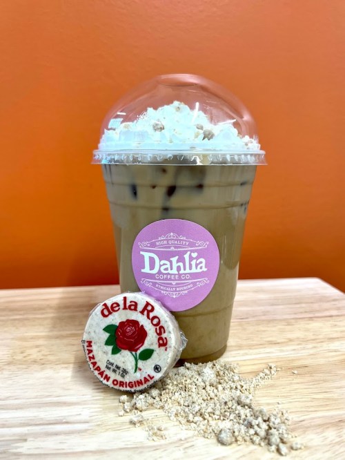 Iced coffee topped with whipped cream, next to a de la Rosa mazapan candy, on a wooden surface with an orange background.
