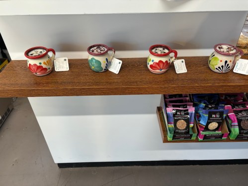 Four colorful ceramic mugs with floral designs displayed on a wooden shelf, alongside bags of snacks below.