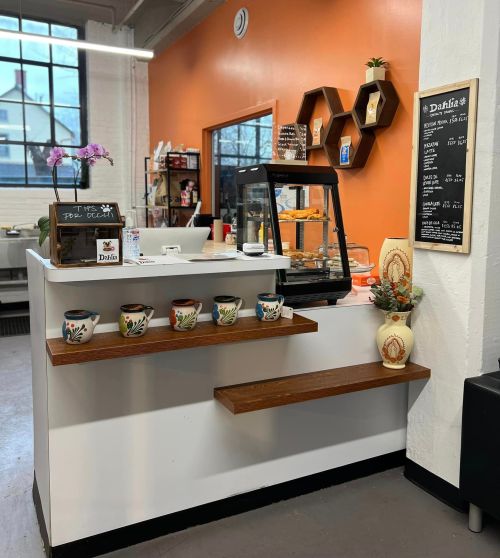 A cozy café counter with baked goods display, decorative shelves, and a chalkboard menu against an orange wall.