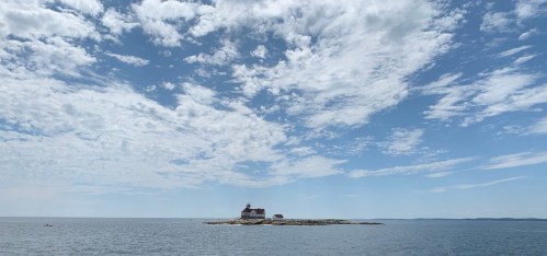 A small lighthouse on a rocky island surrounded by calm waters under a bright, cloudy sky.