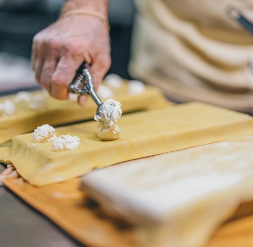 A hand using a scoop to place cheese filling onto a sheet of pasta for making ravioli.