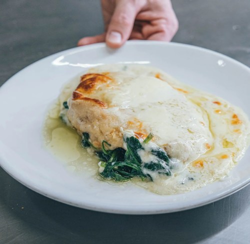 A hand holds a plate with a creamy chicken dish topped with melted cheese and served on a bed of spinach.