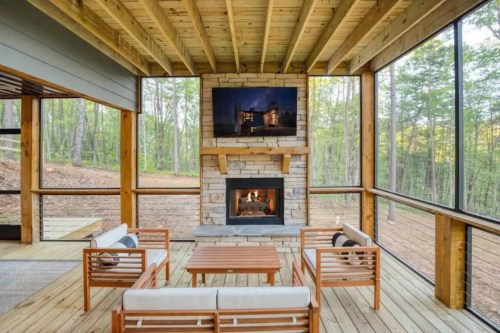Cozy screened porch with wooden furniture, a stone fireplace, and a TV, surrounded by trees and natural light.