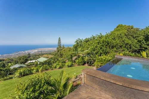 A scenic view from a hillside pool overlooking lush greenery and the ocean in the distance under a clear blue sky.