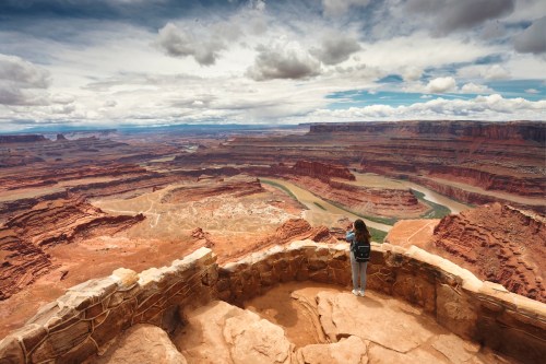 A person stands on a stone overlook, gazing at a vast canyon landscape under a cloudy sky.