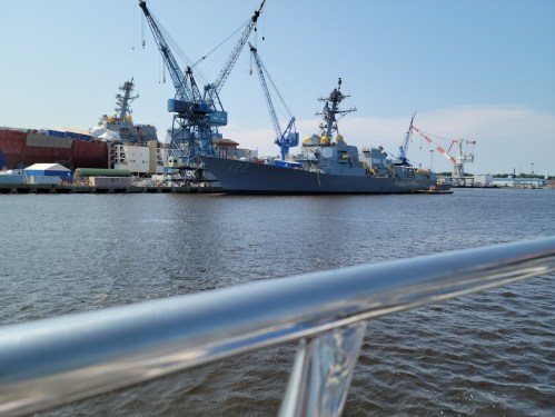A naval ship docked at a shipyard, with cranes and buildings in the background, viewed from a boat on the water.