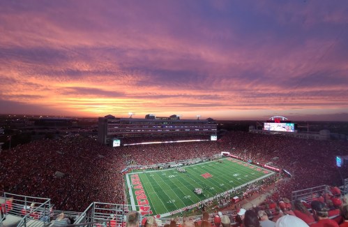 A vibrant sunset over a packed football stadium, with a green field and cheering fans in red attire.