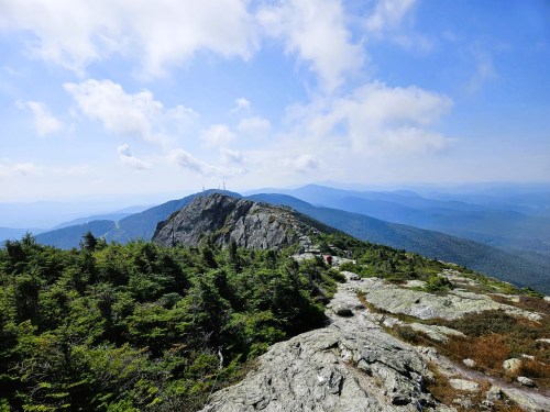 A panoramic view from a mountain peak, featuring rocky terrain, lush greenery, and distant blue mountains under a cloudy sky.