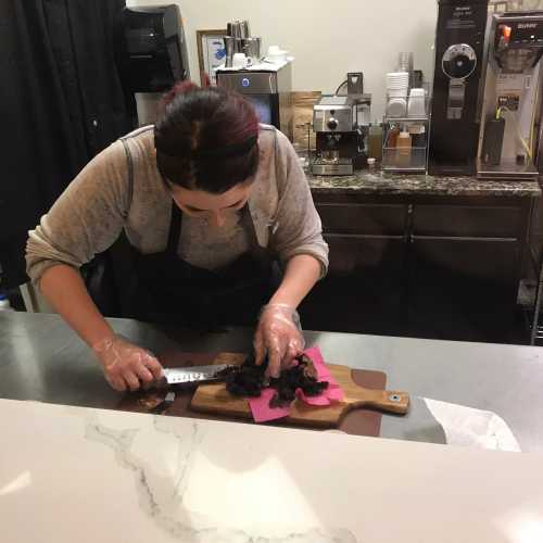 A person in an apron slices food on a wooden cutting board in a cafe kitchen.