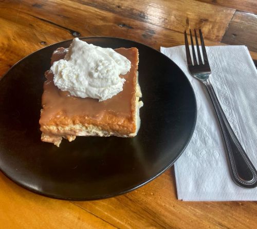 A slice of dessert with creamy topping on a black plate, accompanied by a fork and a napkin on a wooden table.