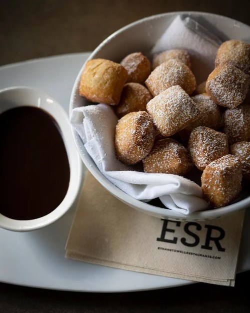 A bowl of powdered sugar-dusted doughnuts with a side of chocolate sauce on a napkin.