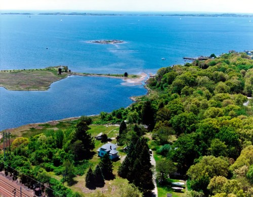Aerial view of a lush green landscape with a blue bay, small islands, and a white house surrounded by trees.