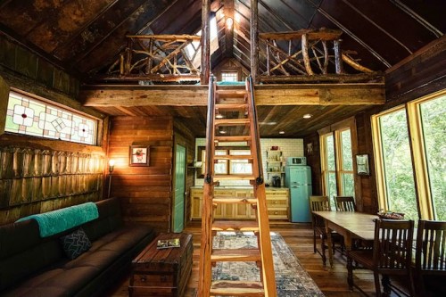 Cozy wooden cabin interior with a loft, kitchen, dining area, and large windows letting in natural light.