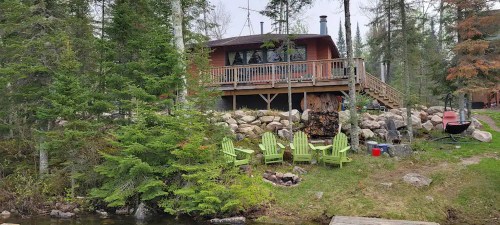 A cozy cabin surrounded by trees, with a deck, stone landscaping, and green chairs by a small water body.