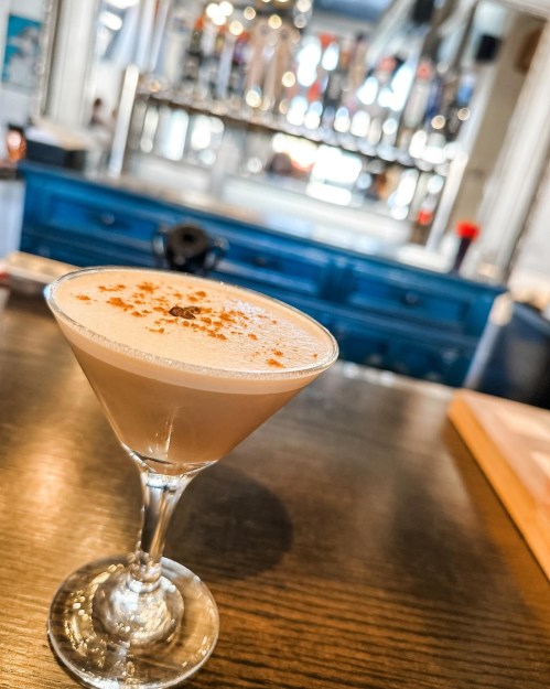 A creamy cocktail in a martini glass, garnished with a sprinkle of cinnamon, on a wooden bar with a blurred background.