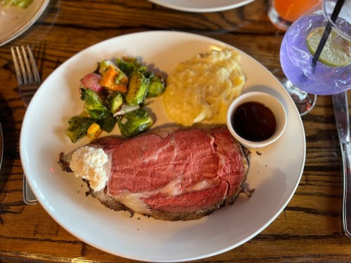 A plate featuring prime rib with horseradish, creamy mashed potatoes, sautéed vegetables, and a dipping sauce.