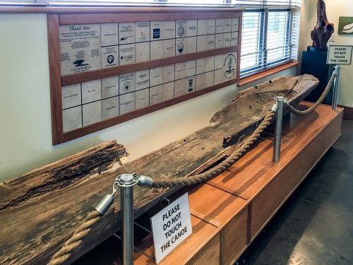 A canoe displayed on a wooden stand with a sign asking not to touch, alongside a wall of plaques.