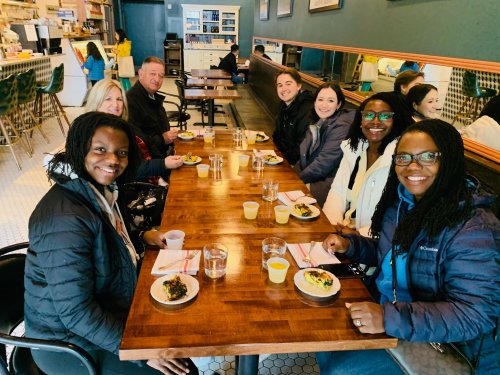 A group of seven people sitting at a wooden table in a cafe, enjoying food and drinks, smiling at the camera.