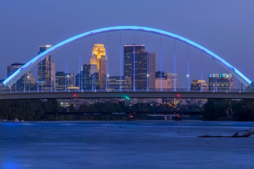 A blue-lit bridge arches over a river, with a city skyline illuminated in the background at dusk.