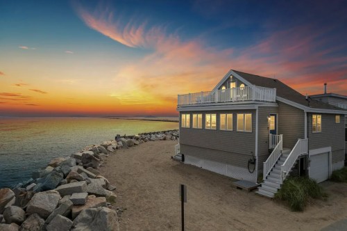 A beach house at sunset, with a rocky shoreline and colorful sky reflecting on the water.