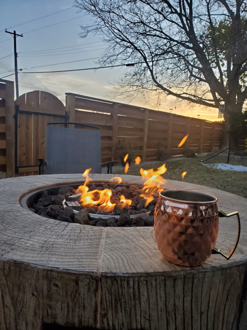 A cozy fire pit with flames, a copper mug nearby, and a sunset sky in the background.