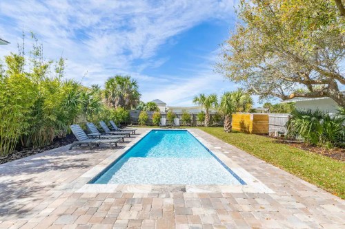 A serene backyard with a sparkling pool, lounge chairs, and lush greenery under a clear blue sky.