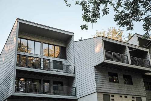 Modern multi-story building with large windows and balconies, surrounded by trees against a clear sky.