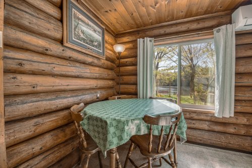 Cozy log cabin interior with a round table, green tablecloth, and a window overlooking trees and a river.