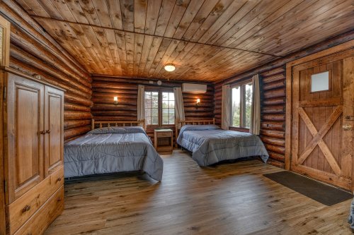 Cozy log cabin interior with two beds, wooden walls, and a window overlooking nature. Warm, rustic atmosphere.
