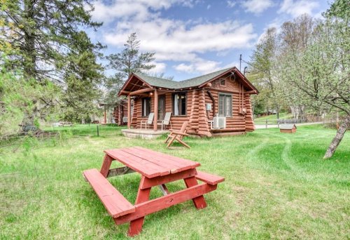 A cozy log cabin surrounded by greenery, featuring a picnic table in front and a clear blue sky above.