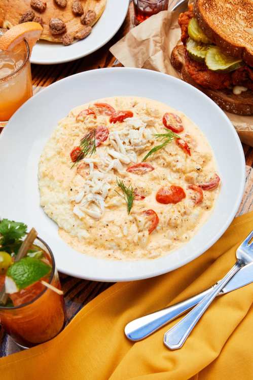A bowl of creamy grits topped with tomatoes and herbs, alongside a drink and a plate of cookies and fried chicken sandwich.
