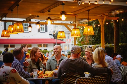 A lively outdoor dining scene with friends enjoying food and drinks under warm lights at a restaurant.