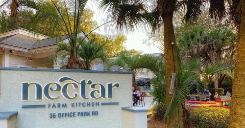 Sign for Nectar Farm Kitchen, surrounded by palm trees, with outdoor seating visible in a warm, inviting setting.